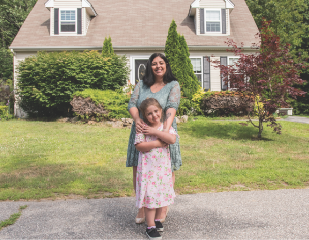 Mother holding her arms across her daughter in front of home.